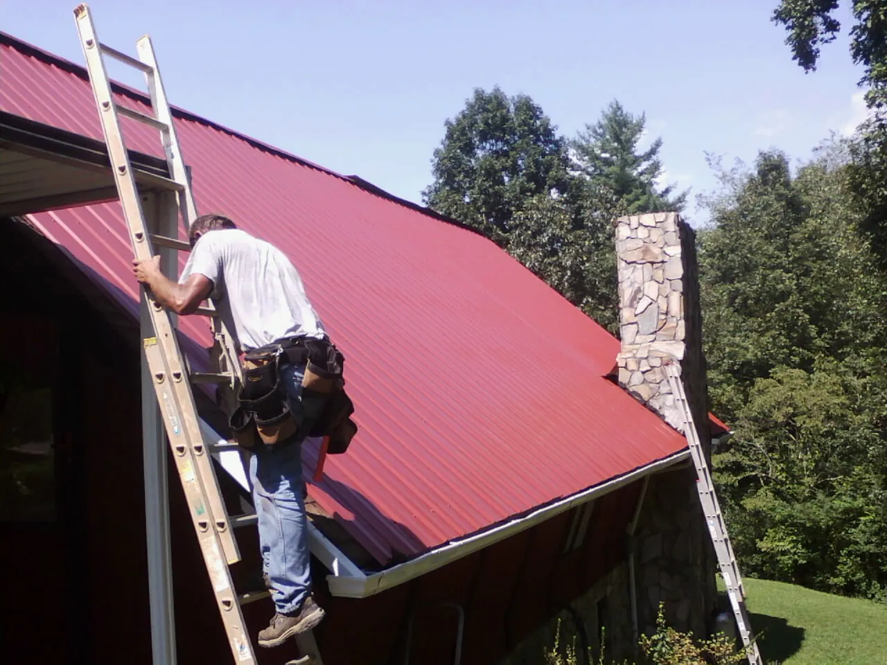 Roofer on a ladder beside a red metal roof and stone chimney — Soffit & Fascia Repair by Roofing Brothers in Coudersport